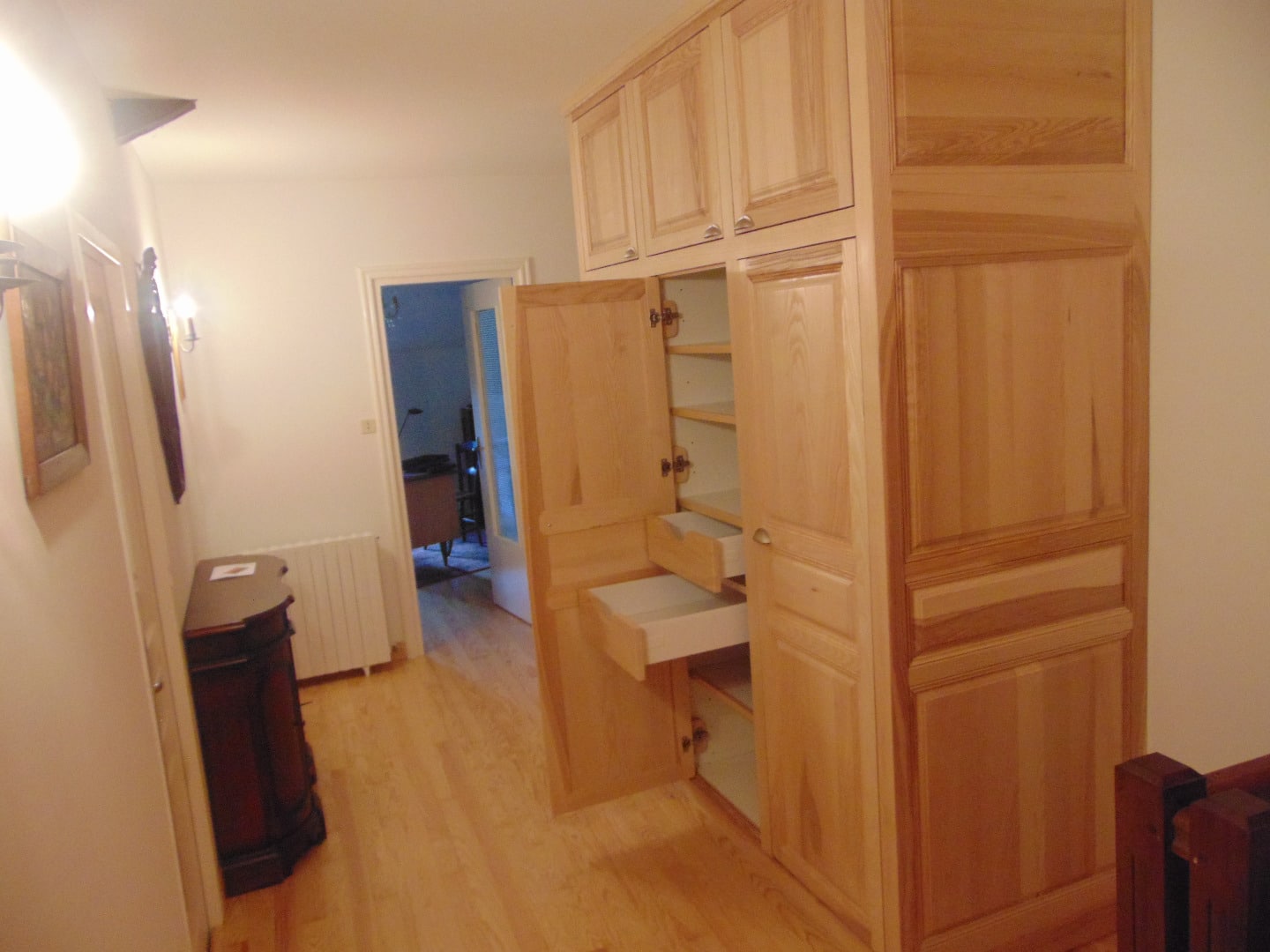 Couloir avec plancher bois clair, une grande armoire en bois naturel ouverte, et une commode foncée contre le mur blanc.
