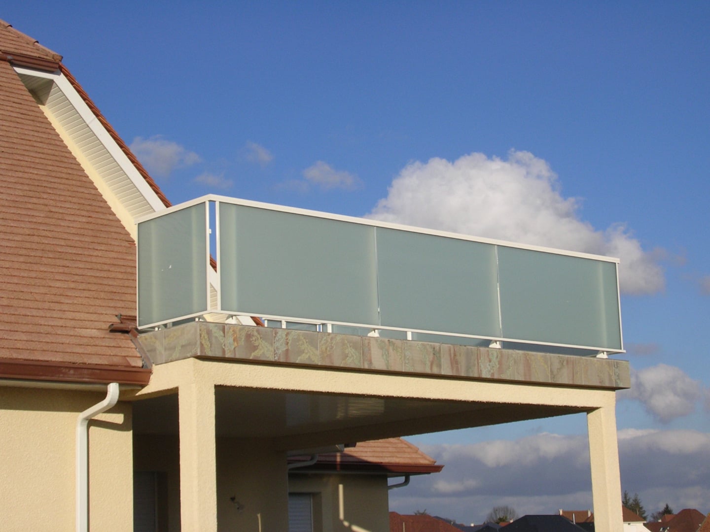 Balcon avec garde-corps moderne en verre dépoli clair et structure blanche, surplombant un toit de tuiles rouges sous un ciel bleu avec nuages.