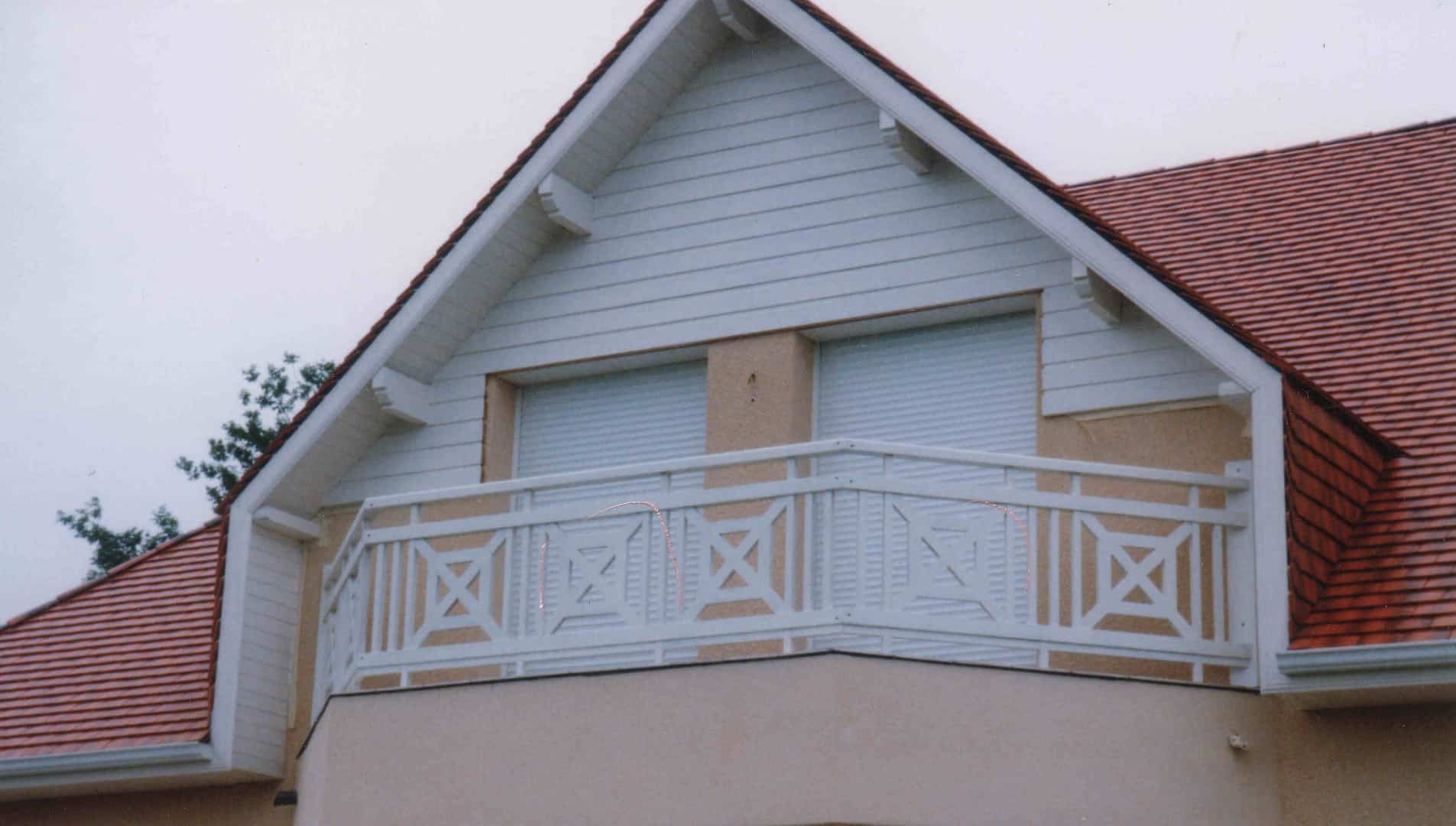 Façade d'une maison avec toit en tuiles rouges et pignon blanc à clin, mettant en vedette un balcon avec balustrade blanche décorative.