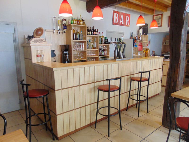 Bar en bois clair avec un comptoir, trois tabourets noirs à assise rouge, des tireuses à bière et une enseigne murale rouge indiquant "BAR".