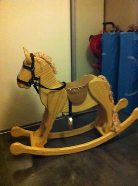 Cheval à bascule artisanal en bois clair avec harnais noir et crinière texturée, posé sur un sol foncé près d'un mur clair.