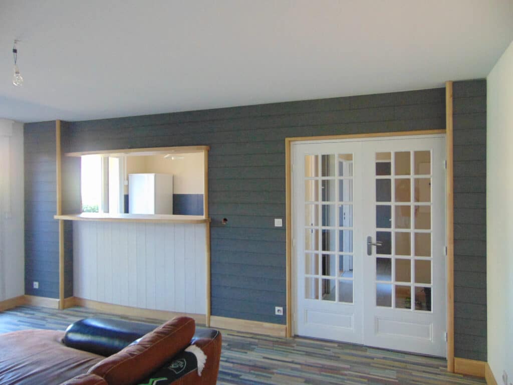 Salon moderne avec mur lambrissé gris foncé, passe-plat en bois clair et portes françaises vitrées blanches menant à un autre couloir.