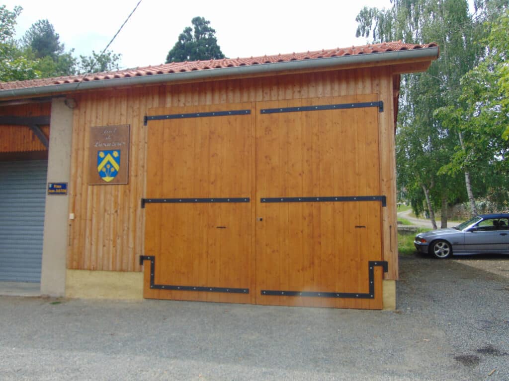 Porte de grange bois blason Larazeres Place Jean JUSTON Grande porte de grange en bois avec ferronnerie noire, panneau blason "Seul de Larazeres", et panneau "Place Jean JUSTON". Voiture garée à droite.