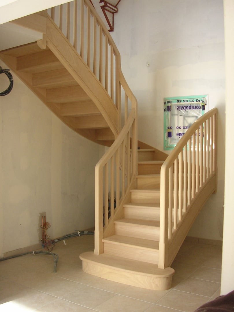 Escalier en bois clair, neuf, courbé, avec balustrades verticales, en cours de chantier (fils apparents). On distingue l'étiquette 'compobale'.