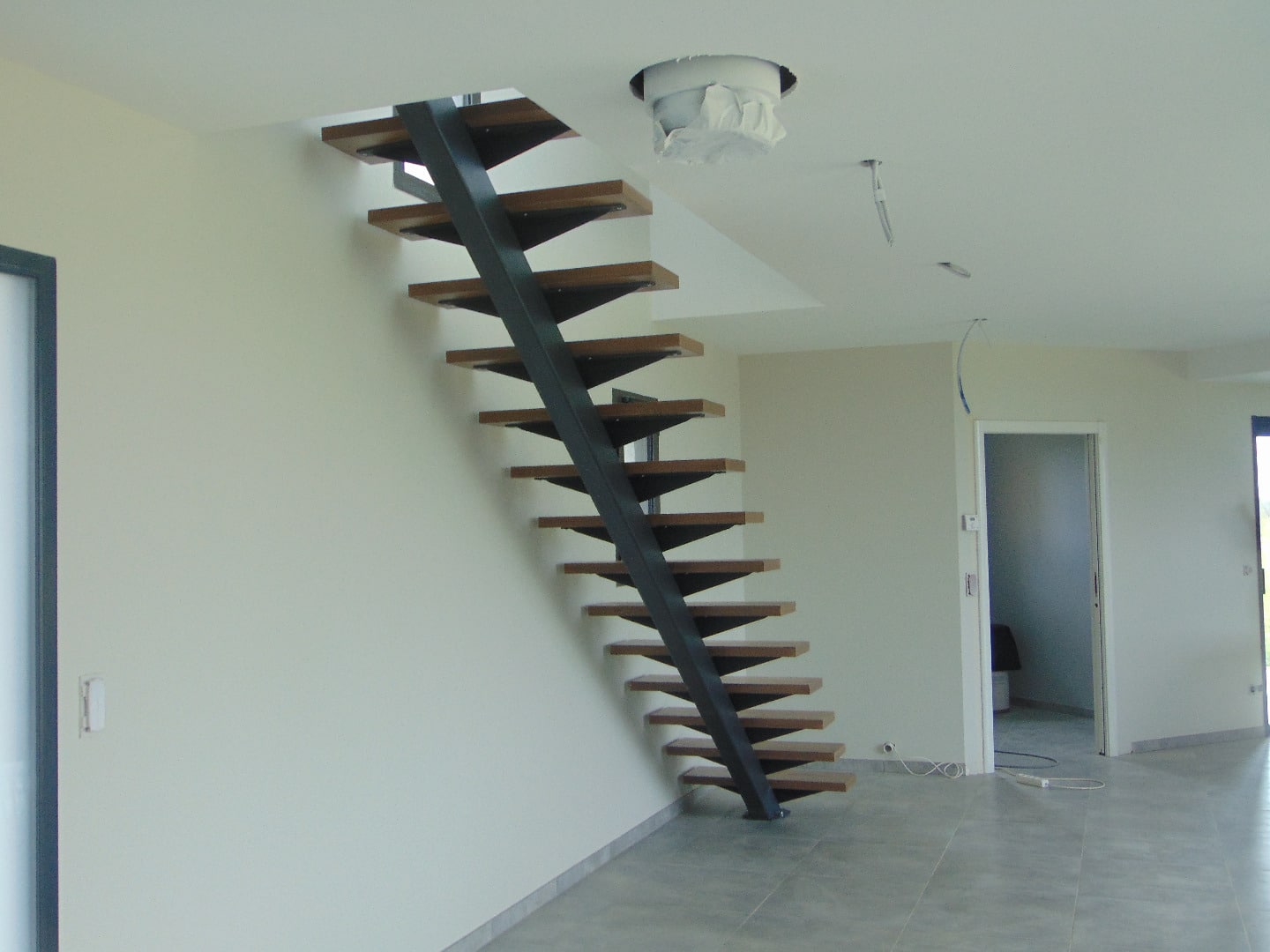 Escalier moderne à limon central noir avec marches en bois flottantes, dans un intérieur clair en cours de finition, câble au plafond.