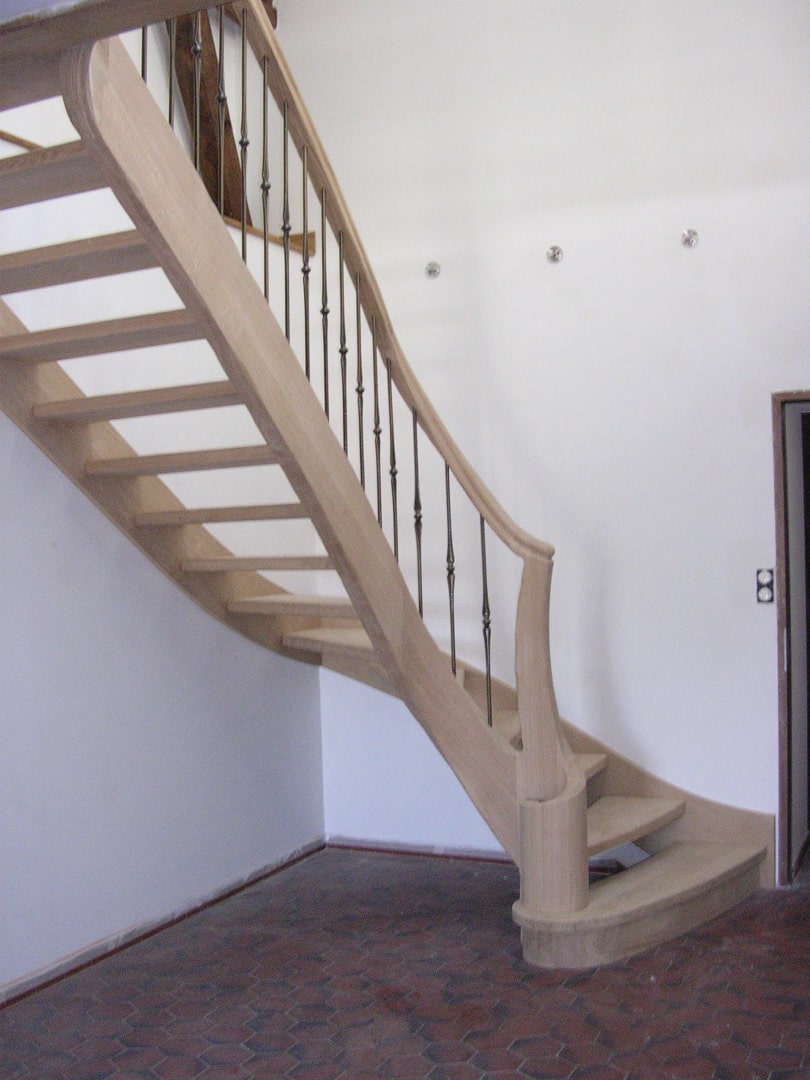 Escalier sur mesure en bois clair, courbé, avec balustres en métal forgé noir, sur un sol en carreaux hexagonaux rouges.
