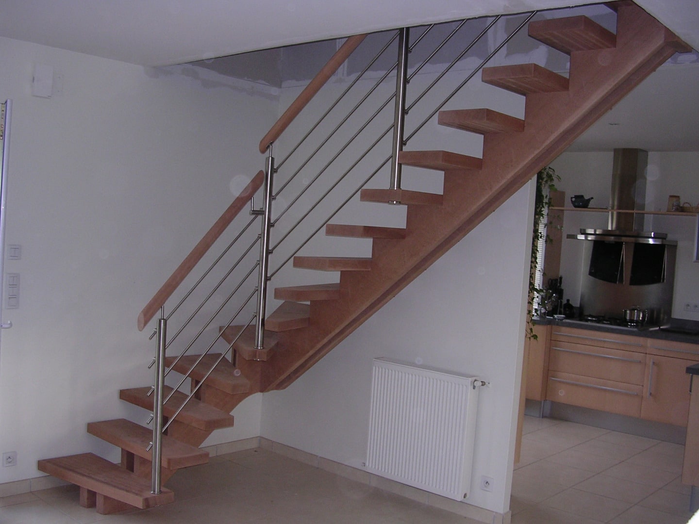 Escalier moderne bois avec limon et garde-corps inox à câbles, près d'un radiateur et ouvrant sur une cuisine équipée.