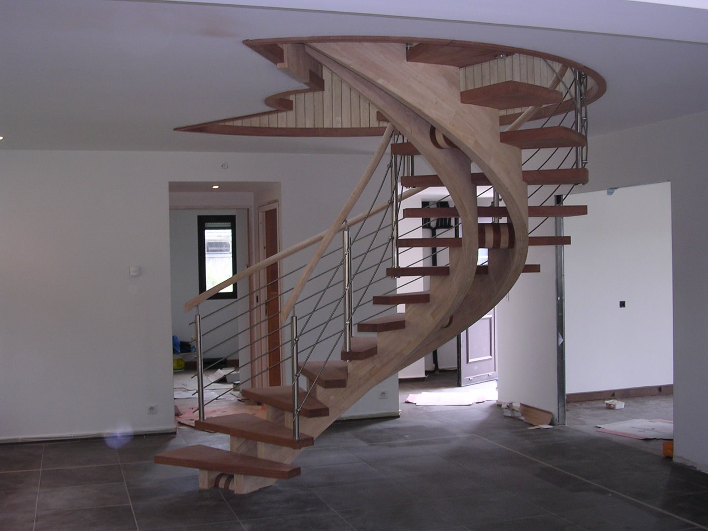 Escalier hélicoïdal moderne en bois clair et bois foncé avec rampe à câbles inox, sous un plafond décoratif sculpté.