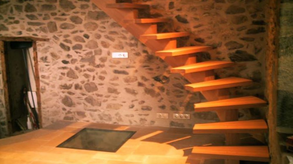 Escalier flottant en bois clair dans une pièce rustique avec murs en pierres apparentes. Une trappe vitrée est au sol. Présence de prises (3x) et d'un boîtier mural (WC).