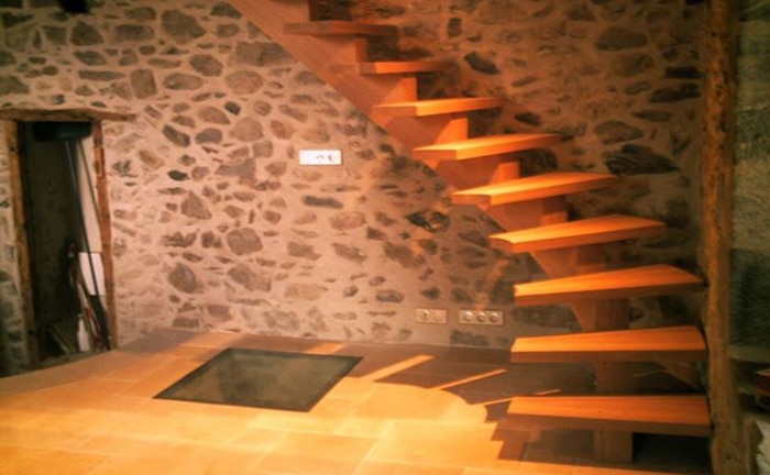Escalier flottant en bois clair moderne devant un mur de pierres rustiques, avec une trappe en verre au sol. Prises électriques visibles.