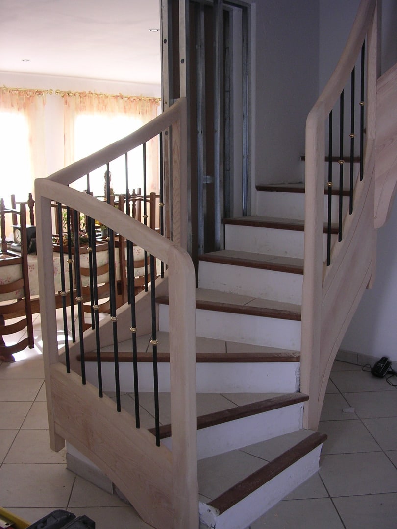 Escalier en bois clair en cours de finition, avec balustres noirs et dorés, près d'une zone de construction intérieure.