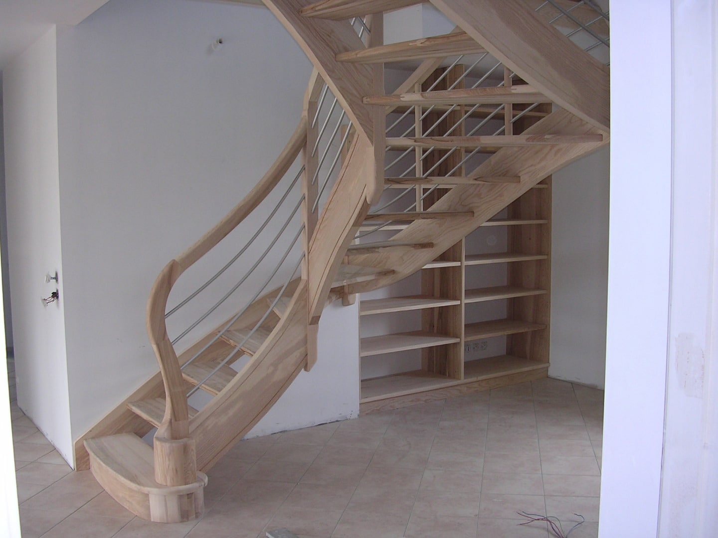 Escalier droit en bois clair avec garde-corps à lisses métalliques horizontales, intégrant des étagères sous-pente.