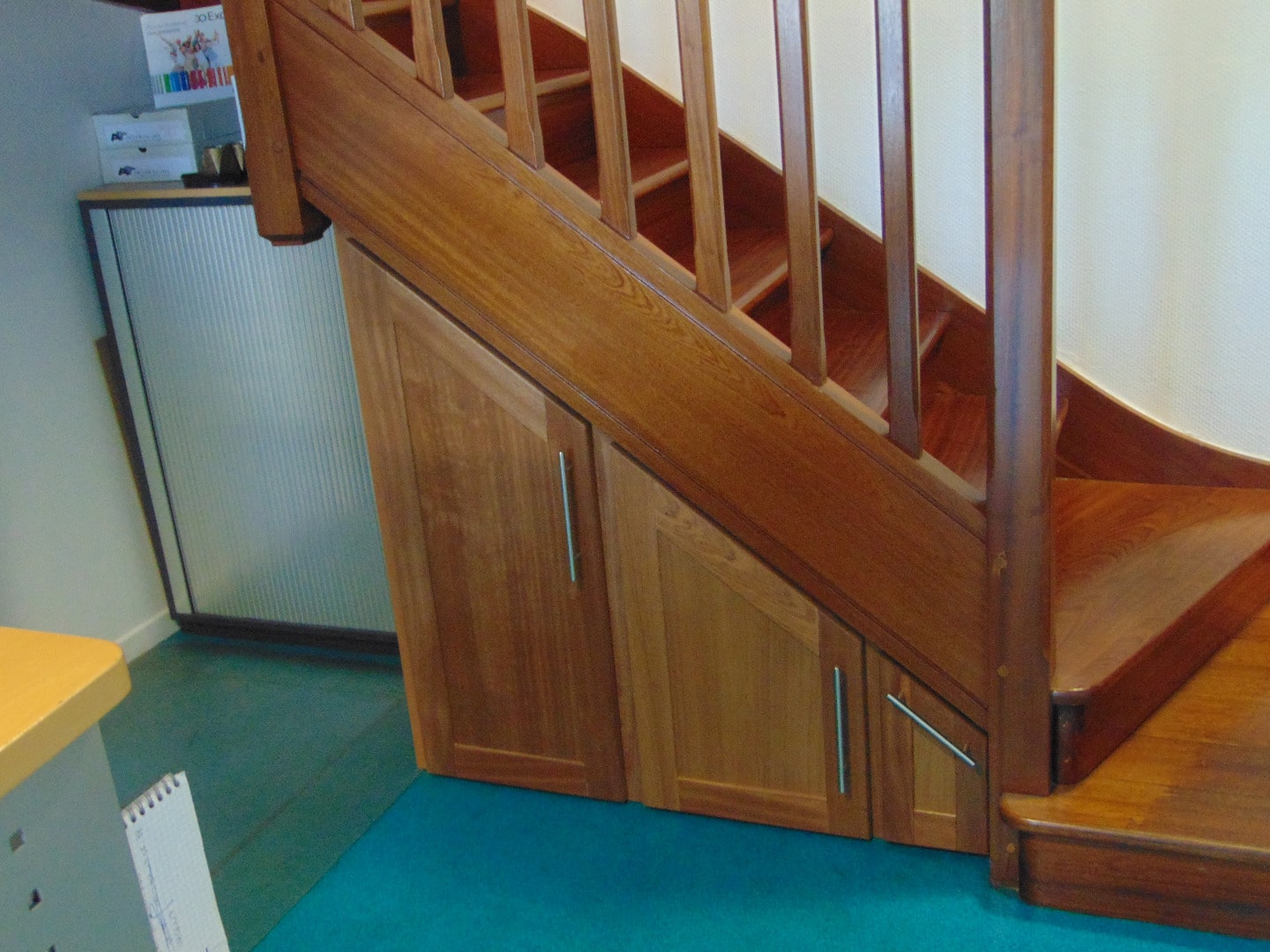 Escalier en bois verni avec rangement sous les marches et une rampe à balustres verticaux. Sol turquoise.