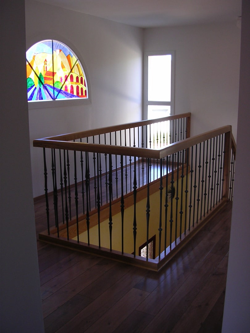 Hall supérieur avec plancher bois foncé et balustrade en fer forgé et bois menant à un escalier, éclairé par vitrail cintré.