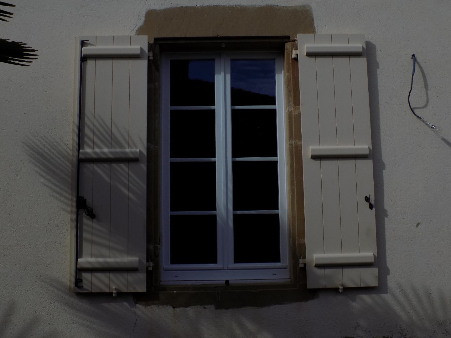 Fenêtre rectangulaire avec volets extérieurs blancs ouverts sur une façade claire, ombres de feuilles de palmier projetées.