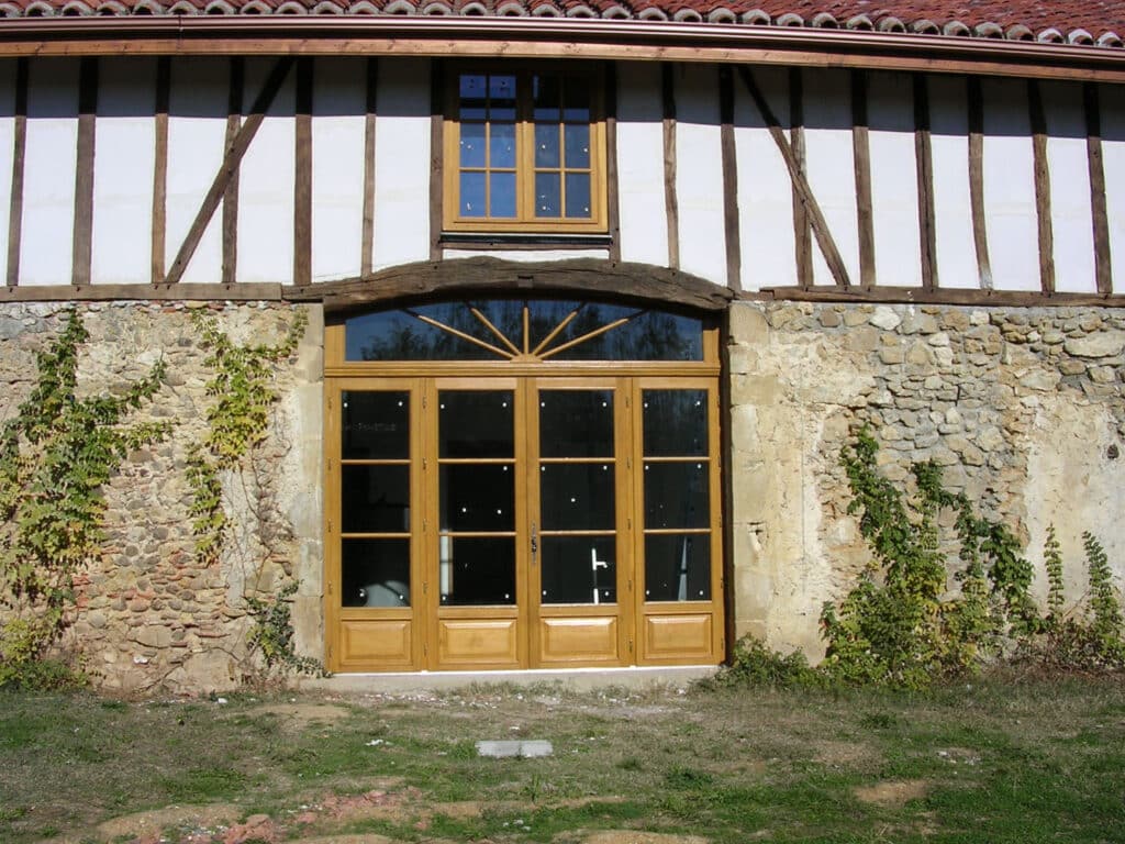 Façade de grange ancienne en colombages à colombages blancs et pierre, avec une grande porte-fenêtre en bois clair et une petite fenêtre à l'étage.