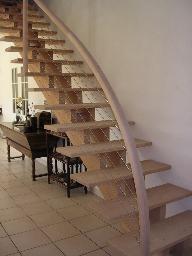 Escalier moderne à marches flottantes en bois clair, avec rampe courbée en bois et garde-corps à câbles métalliques fins.
