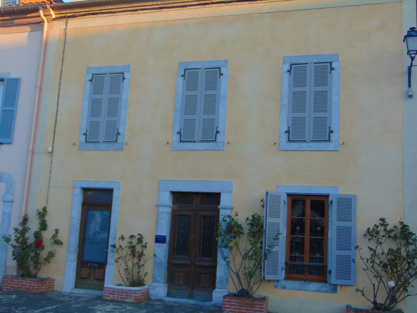 Façade jaune ocre d'une maison avec trois fenêtres à volets gris-bleu à l'étage et deux portes d'entrée au rez-de-chaussée, pots de fleurs.