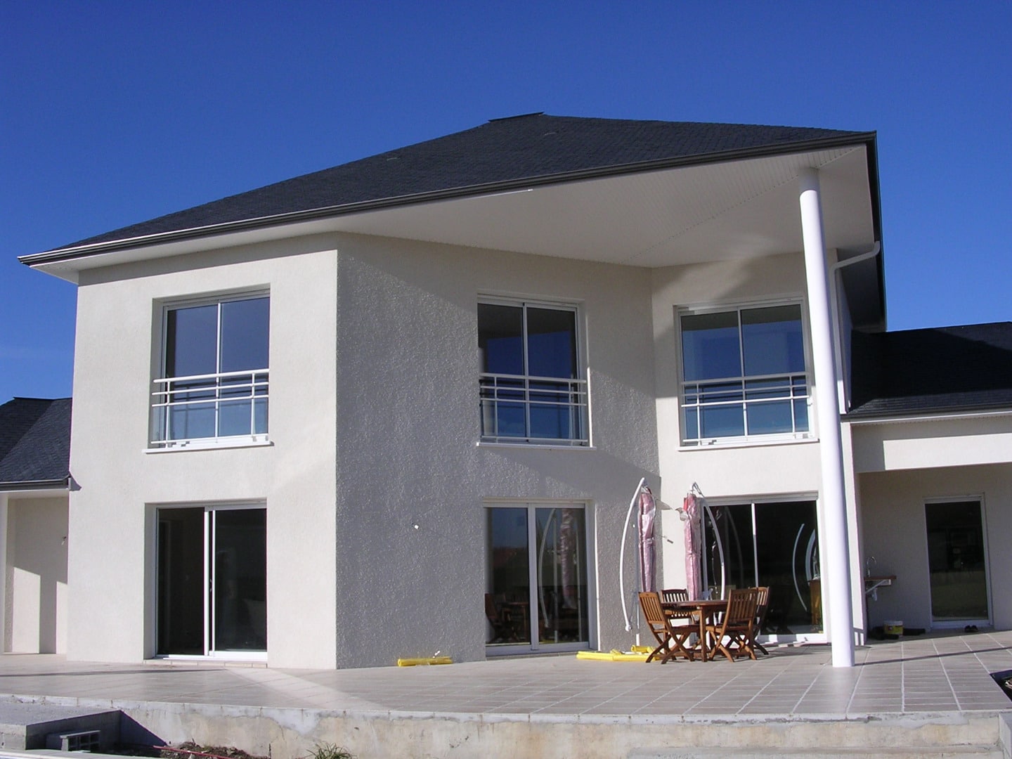 Maison contemporaine à étage avec enduit texturé blanc, toit sombre et large terrasse dallée sous avancée de toit soutenue par un poteau blanc.