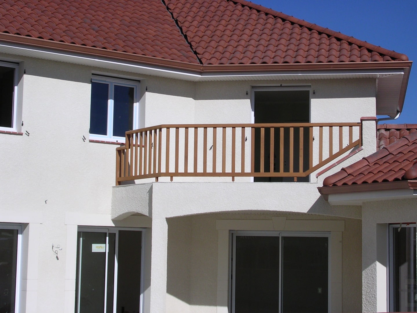 Façade d'une maison neuve avec crépi blanc, toit en tuiles rouges, et un balcon avec garde-corps en bois brun.
