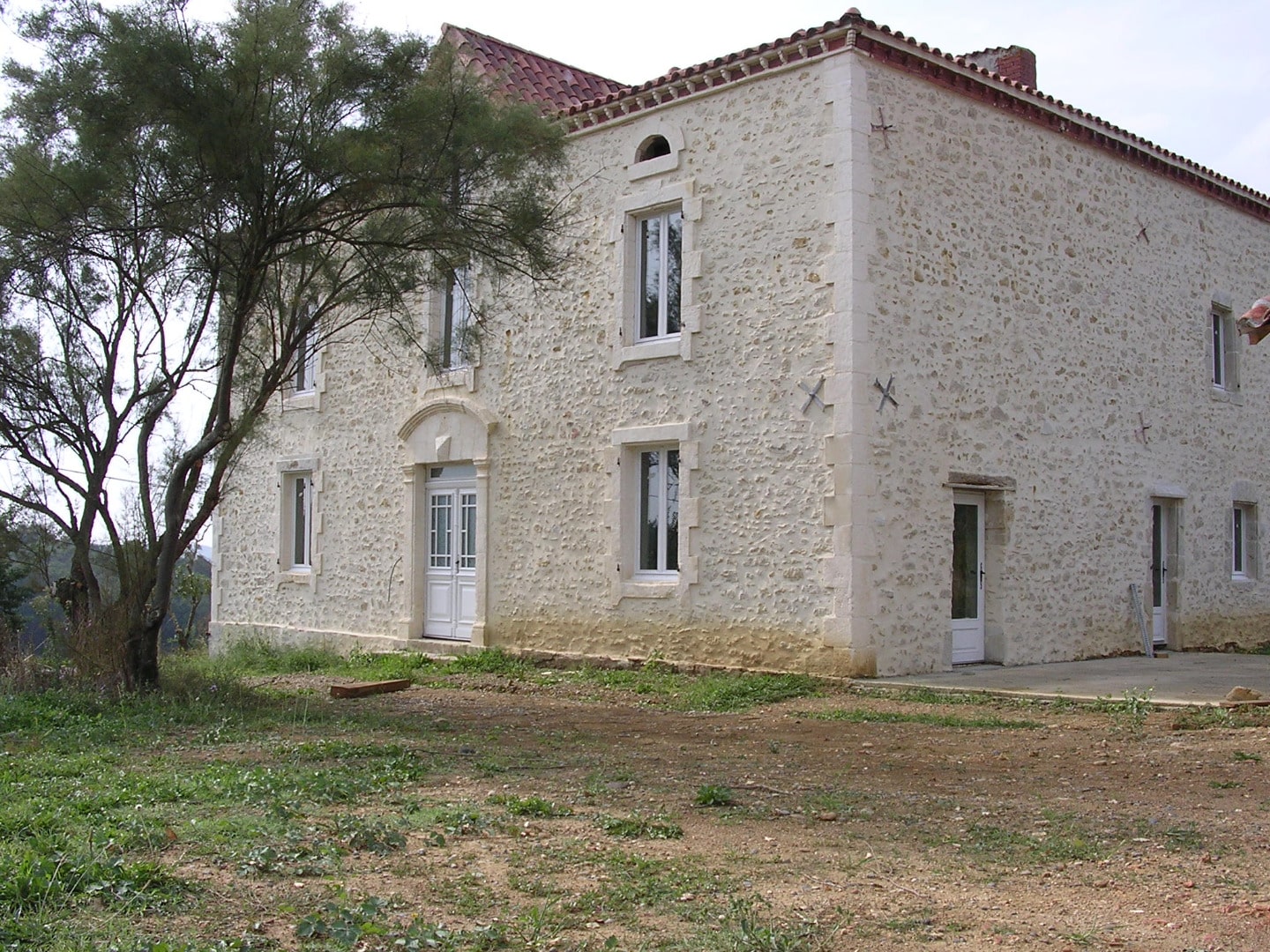 Grande maison en pierre de taille et moellons, avec toiture en tuiles rouges, devant un arbre touffu et un terrain terreux.
