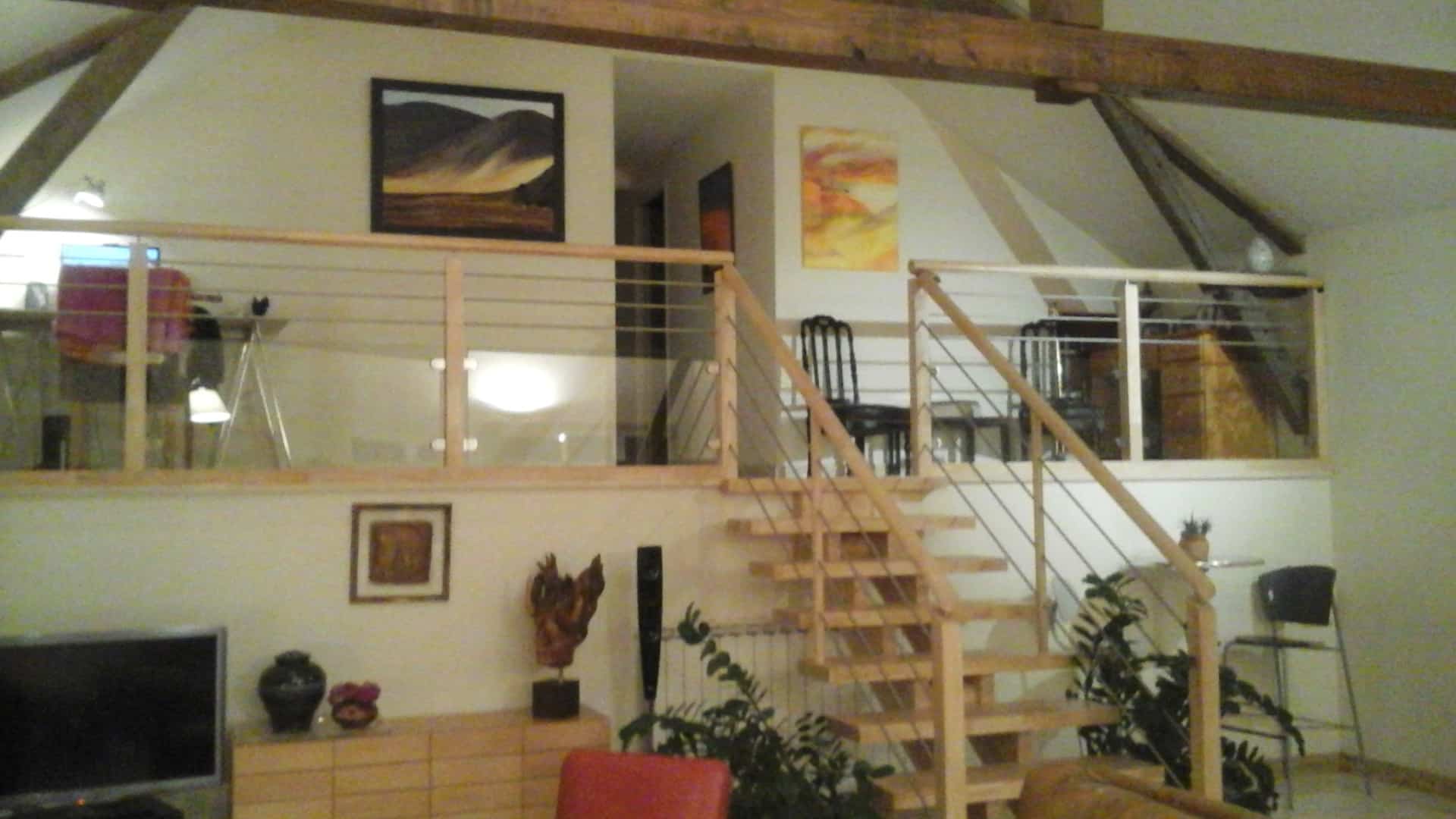 Intérieur moderne avec mezzanine et escalier ouvert en bois clair, garde-corps câble inox et verre, surplombant un meuble bas TV.