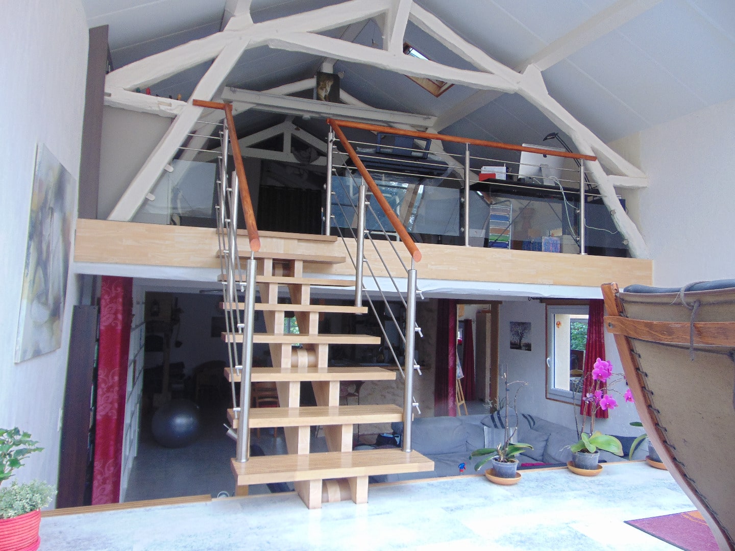Escalier moderne avec marches en bois clair et garde-corps acier/bois menant à une mezzanine bureau sous charpente blanche.