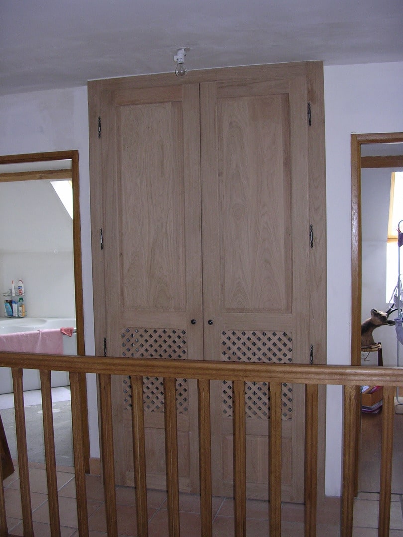 Rambarde en bois au premier plan devant un grand placard double en chêne clair avec aérations losangées. À gauche, accès à une salle de bain.