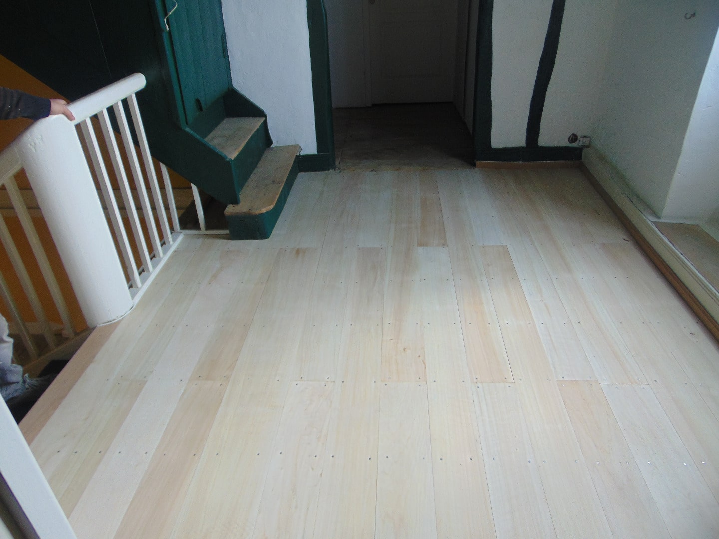 Pose de plancher en bois clair, cloué, près d'une balustrade blanche et d'un escalier peint en vert foncé.