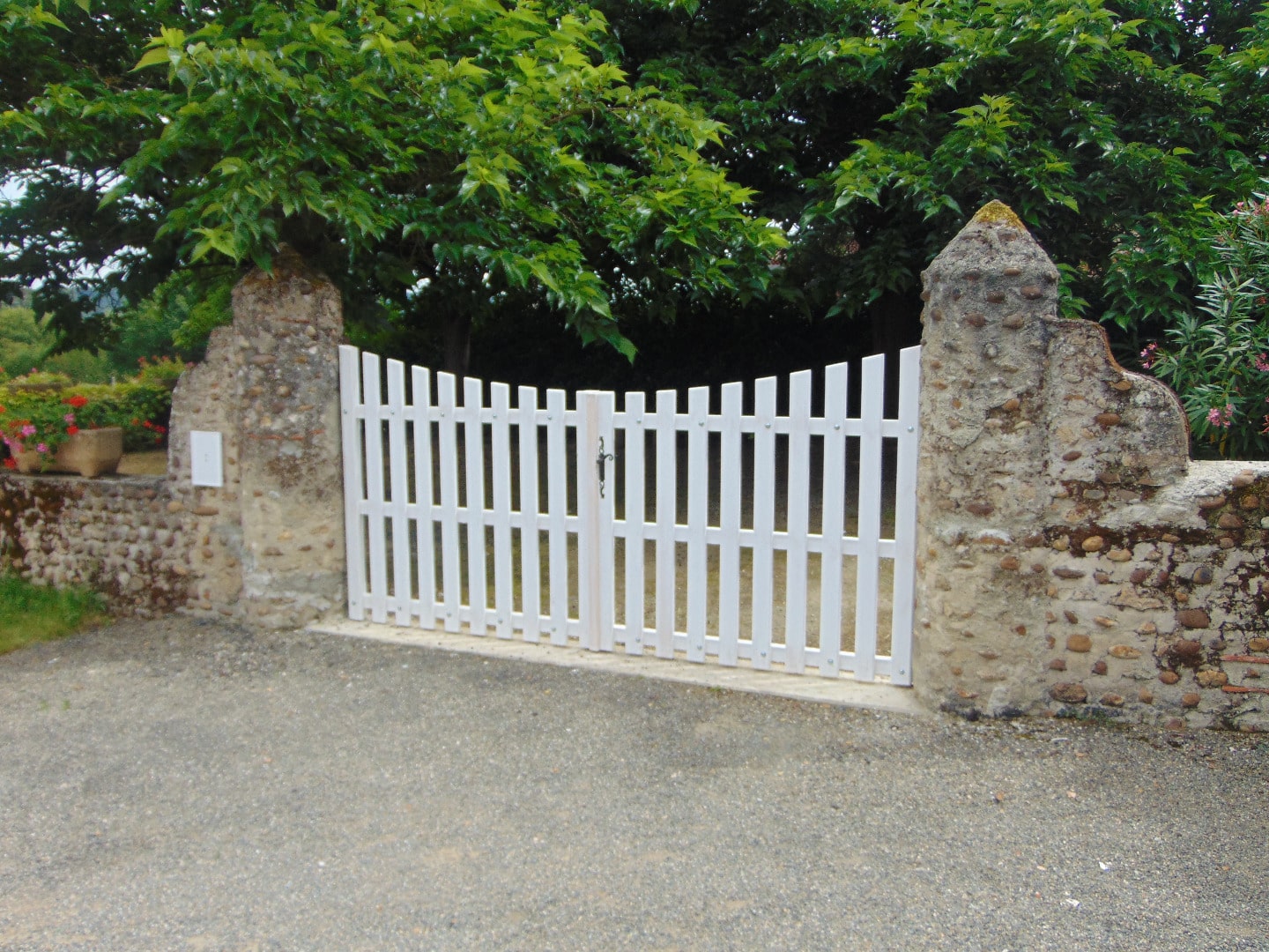 Portail blanc à claire-voie entre deux piliers en pierre brute, devant une allée gravillonnée et une végétation luxuriante.