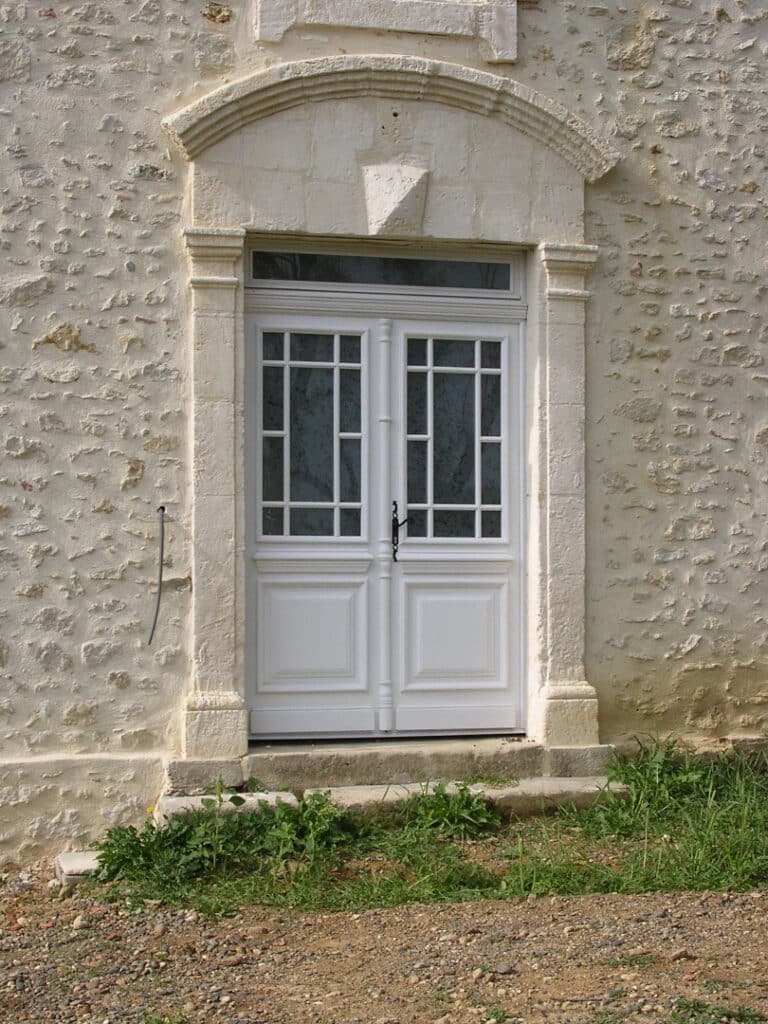 Porte d'entrée double blanche à petits carreaux, encadrée de pierre calcaire travaillée sur un mur rustique, avec marches en pierre et végétation.