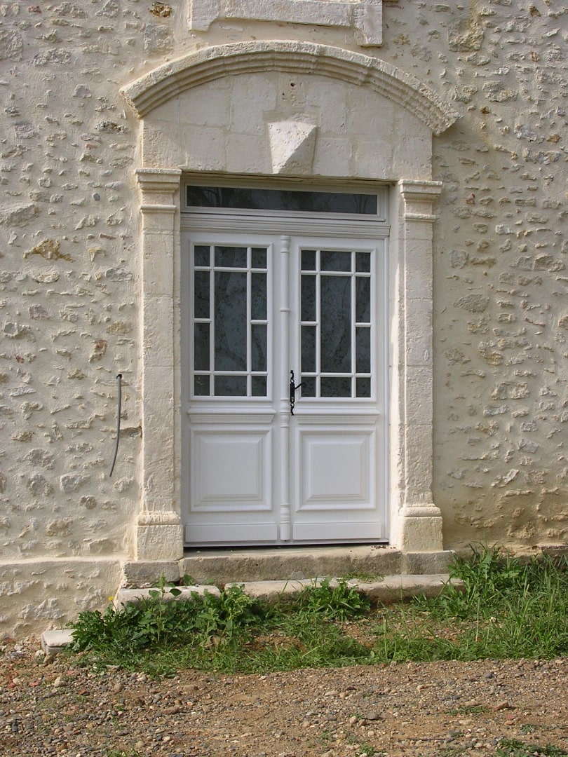 Porte d'entrée double blanche à petits carreaux, encadrée de pierre calcaire travaillée sur un mur rustique, avec marches en pierre et végétation.