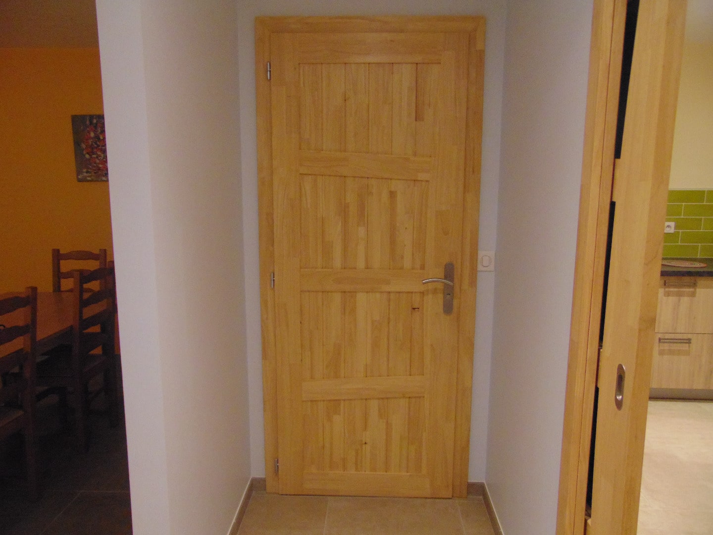 Intérieur avec une porte battante en bois clair dans un couloir, menant à une cuisine verte et une salle à manger orange.