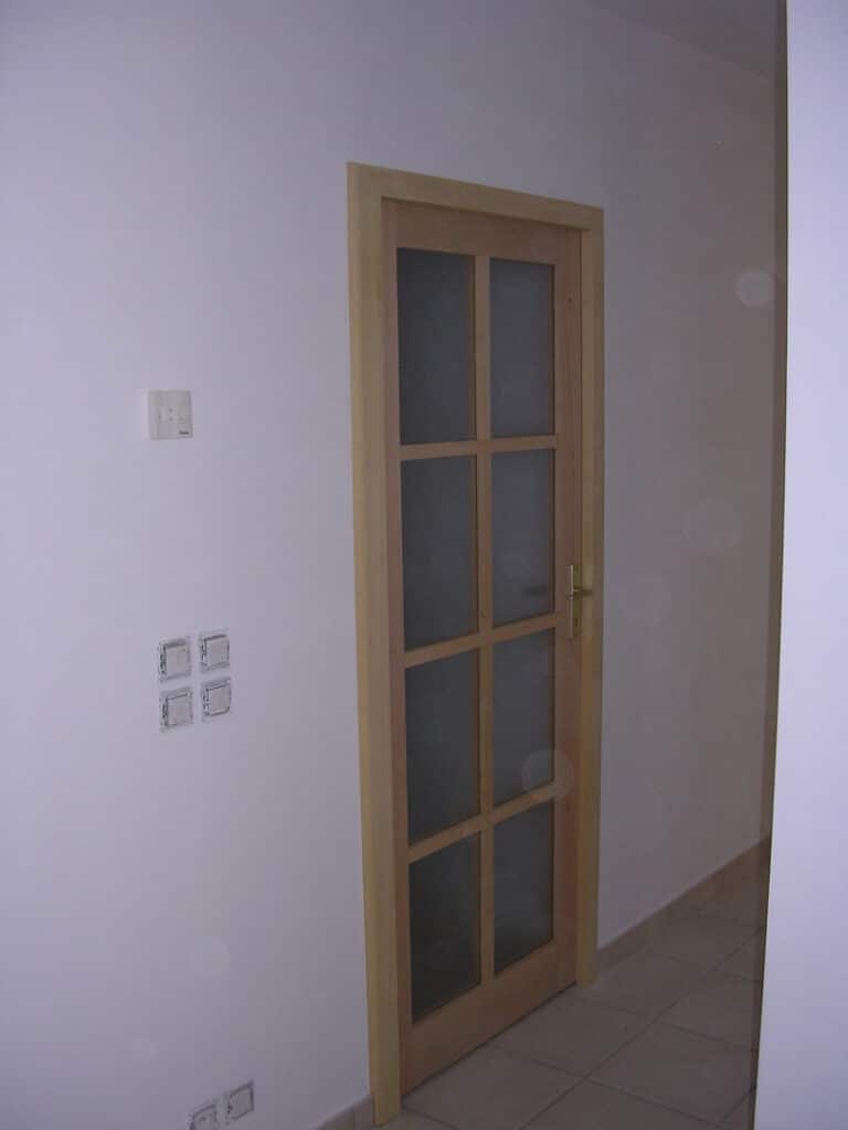 Porte intérieure à cadre bois clair avec six panneaux de verre dépoli et quincaillerie dorée, installée dans un couloir aux murs blancs et sol carrelé.