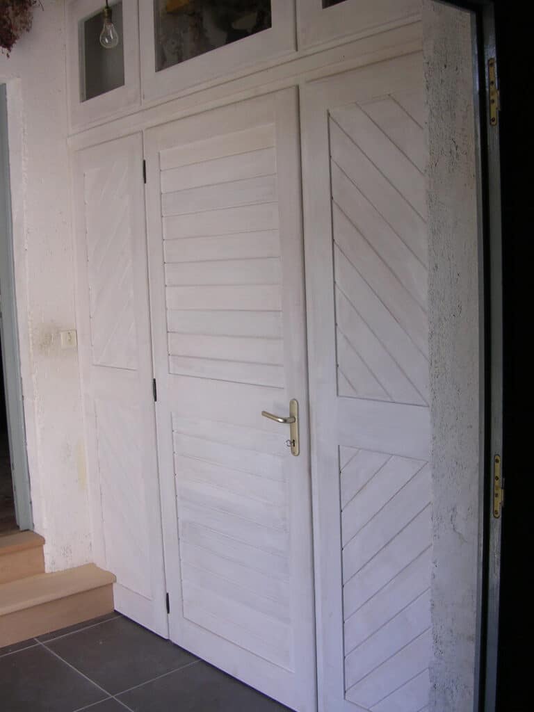 Porte d'entrée double blanche à volets avec panneaux à chevrons et petites fenêtres supérieures, poignée laiton, sol carrelé foncé.