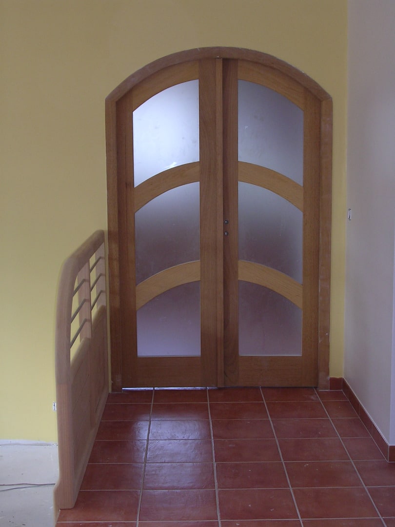 Porte double en bois cintrée avec vitrage dépoli et traverses courbes, sur sol en carreaux de terre cuite.