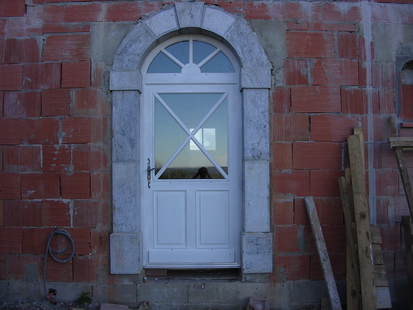 Porte d'entrée blanche neuve avec imposte vitrée arquée, encadrée de pierre grise sur mur en briques rouges de chantier.