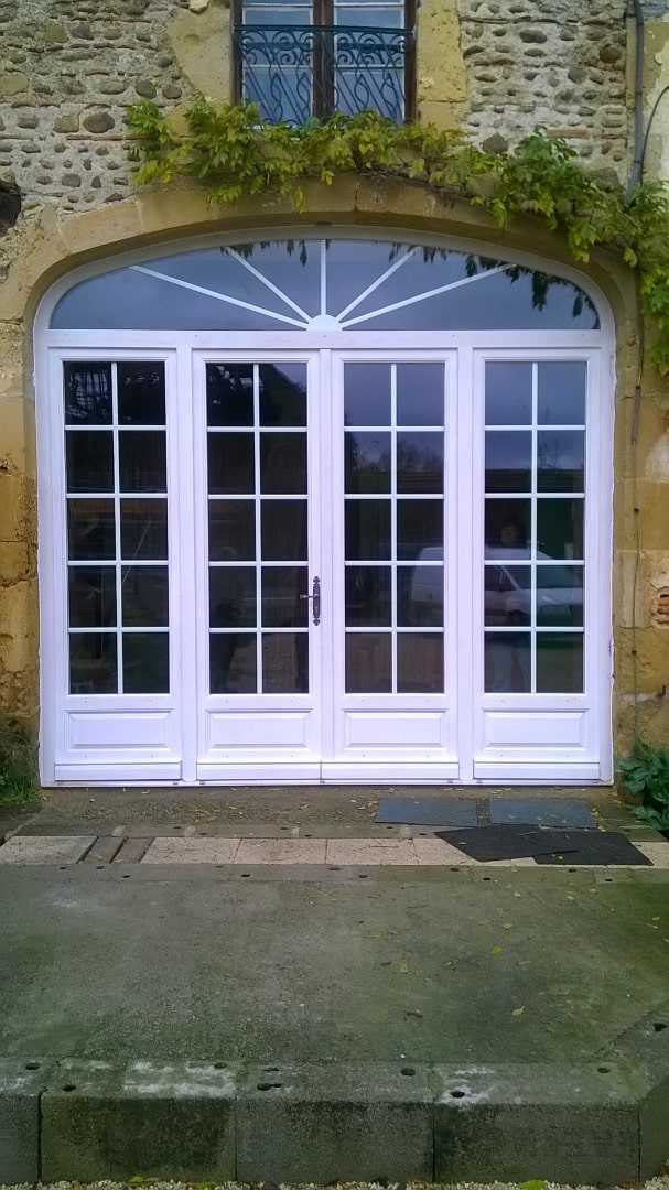 Grande porte-fenêtre cintrée blanche à quatre vantaux, encadrée de pierre, avec petits carreaux et imposte en éventail au-dessus.