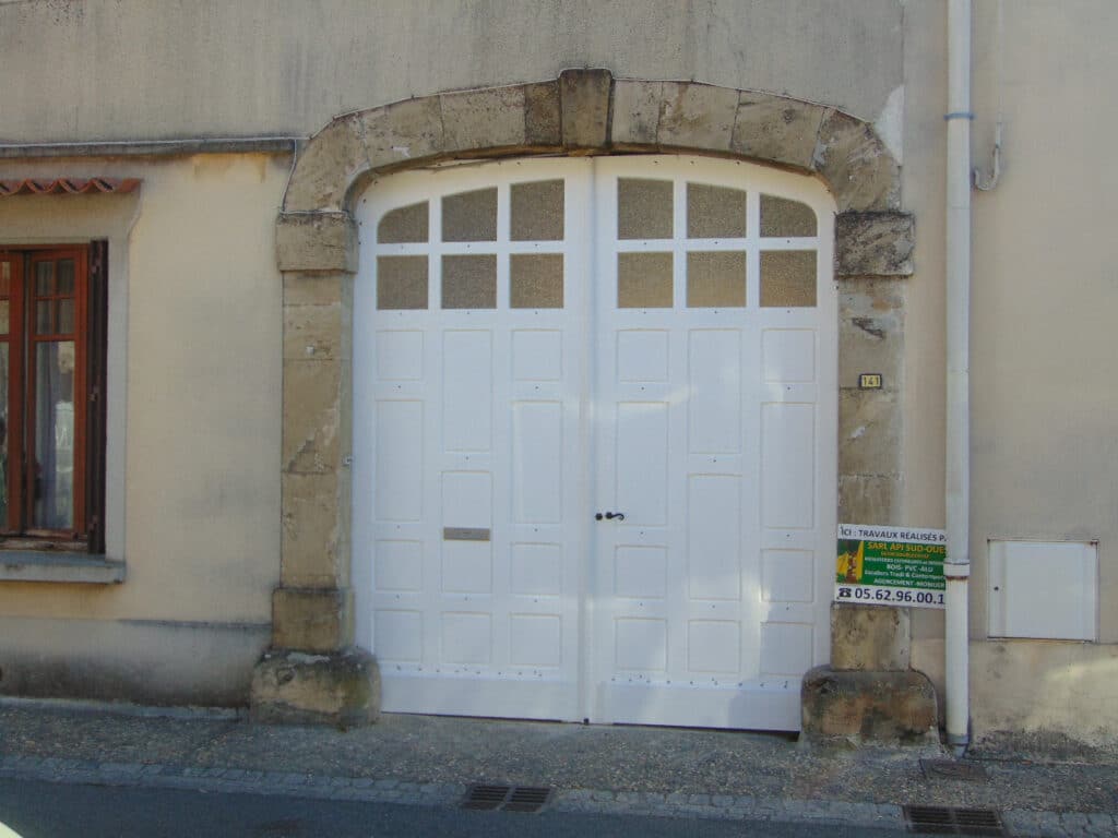 Porte de garage double blanche sous une arche en pierre, façade beige, affiche "SARL API SUD-OUEST 05.62.96.00.1".