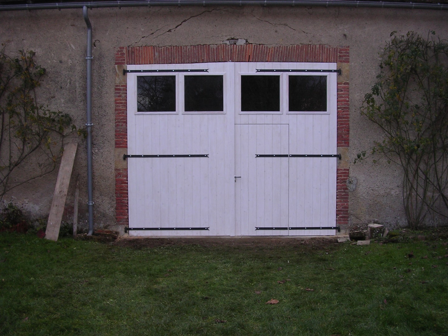 Double porte de garage blanche neuve avec fenêtres, encadrée de briques rouges, devant un mur de pierre et un jardin en herbe.