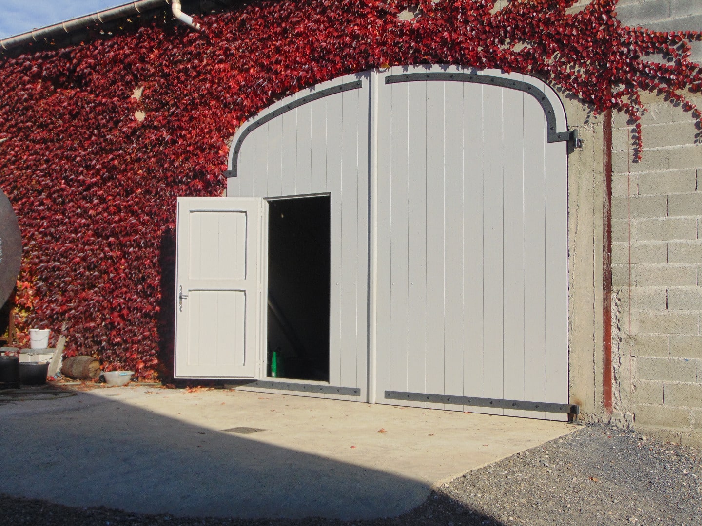 Grande porte de grange grise à deux vantaux avec une petite porte ouverte, devant un mur couvert de vigne vierge rouge flamboyant.