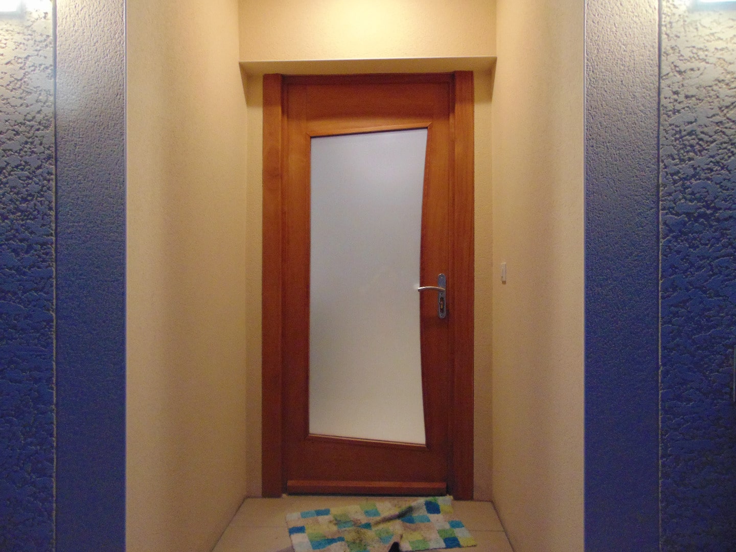 Entrée intérieure avec porte en bois clair et panneau vitré dépoli, encadrée de murs beiges et colonnes texturées bleu foncé.