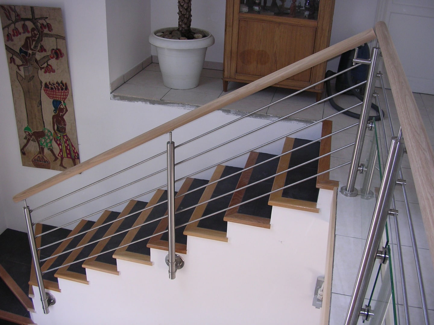 Rambarde moderne d'escalier avec main courante bois clair, câbles inox horizontaux et poteaux chromés. Peinture murale africaine visible.