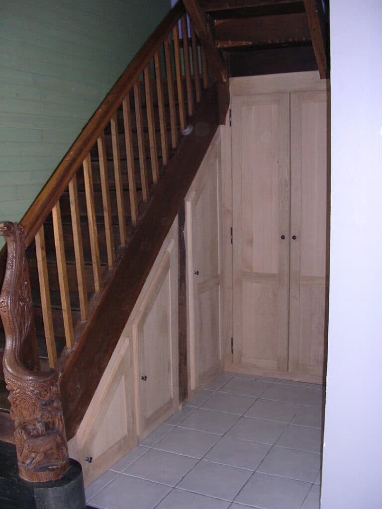 Escalier en bois sombre avec balustre sculpté et rangements intégrés sous les marches, sol carrelé clair.