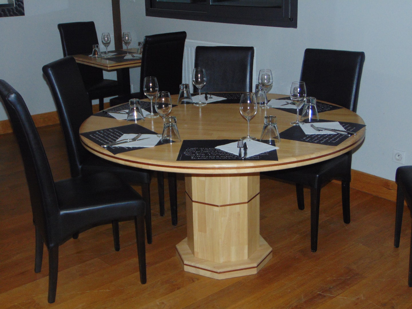 Table ronde en bois clair dressée pour un dîner avec des chaises noires en similicuir, couverts et verres à vin sur sets noirs.