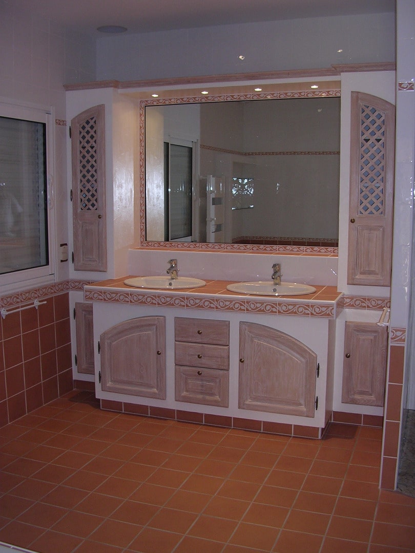 Salle de bain rustique avec meuble double vasque en bois clair, carrelage terre cuite au sol et miroir encadré.