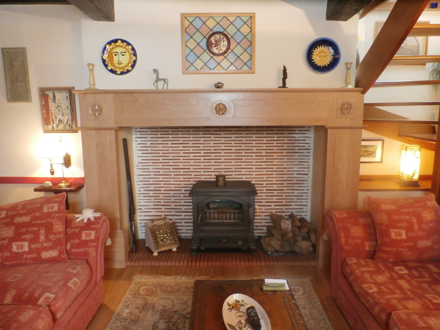 Salon cosy avec cheminée à poêle en fonte, manteau en bois clair sculpté, décorations murales solaires et escalier en bois à droite.