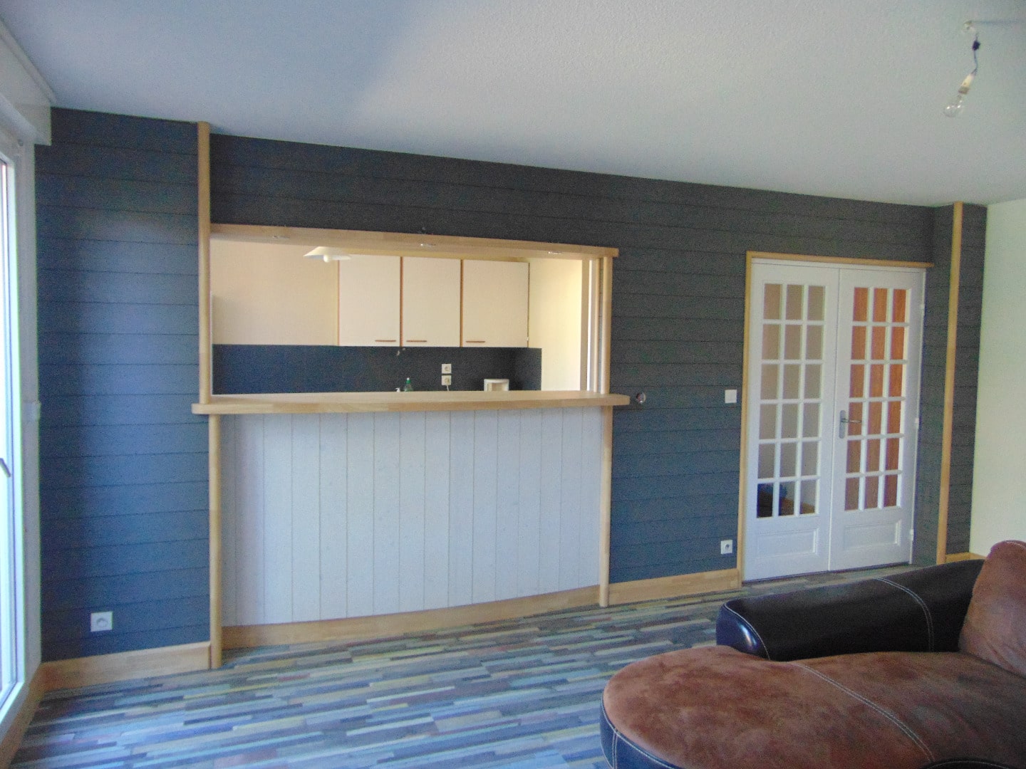 Salon moderne avec mur lambrissé gris foncé, passe-bar en bois clair séparant du séjour, et portes-fenêtres blanches à petits carreaux.